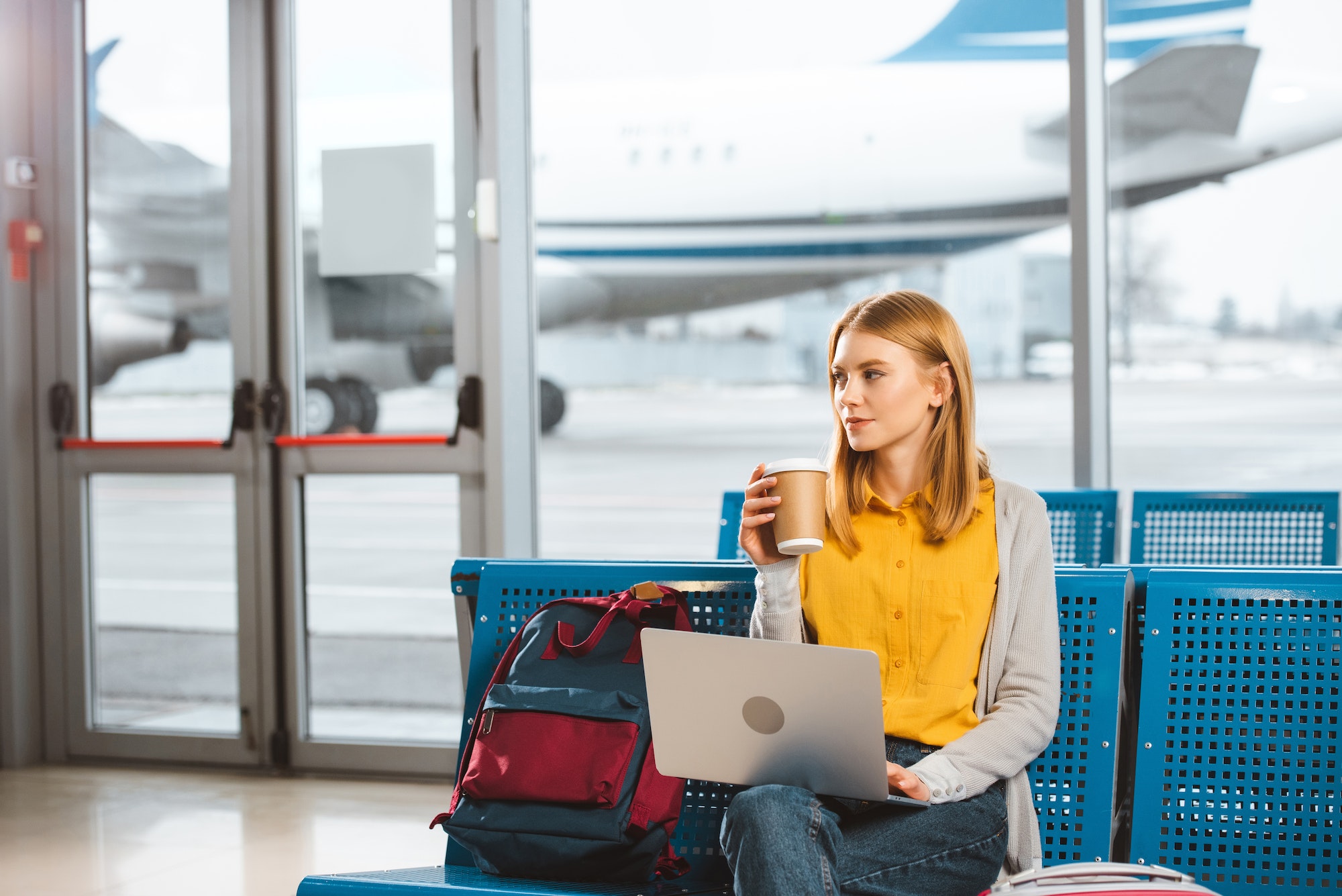 beautiful woman sitting with laptop and holding disposable cup in hand in airport
